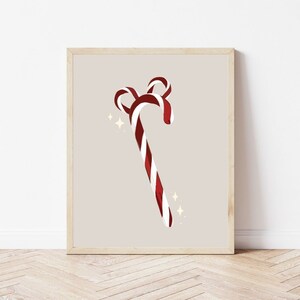 Op de afbeelding: Een ingelijste print met een gestileerde zuurstok met rood-witte strepen. De zuurstok heeft een gebogen top en staat tegen een neutrale achtergrond met sprankelende accenten, wat een feestelijk thema oproept.