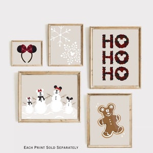 May include: A collection of framed Christmas-themed prints. The prints feature a Minnie Mouse ear headband, snowflakes, the word "HO HO HO", snowmen, and a gingerbread man. Each print has a beige background and a wooden frame.