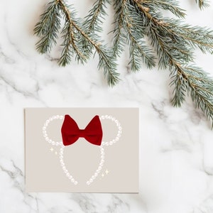 May include: A greeting card featuring a red bow and pearl-like beads arranged in the shape of a mouse head. The card has a light beige background with sparkling accents. A sprig of evergreen is in the upper right corner.