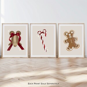 Op de afbeelding: Drie ingelijste kunstprints met kerstthema-illustraties. De prints tonen een gouden bel met een rood lint, een rood-witte zuurstok en een peperkoekmannetje. Elke print staat op een beige achtergrond.