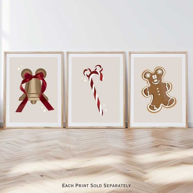 Op de afbeelding: Drie ingelijste kunstprints met kerstthema-illustraties. De prints tonen een gouden bel met een rood lint, een rood-witte zuurstok en een peperkoekmannetje. Elke print staat op een beige achtergrond.