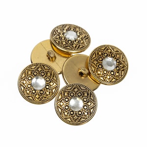 May include: A selection of decorative, gold-toned buttons, each with a clear, central gem. The buttons have an embossed, patterned design around the gem and a loop on the reverse for sewing. The buttons are circular and have a vintage appearance.