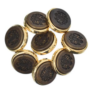 Puede incluir: Un conjunto de ocho botones redondos con un centro marrón oscuro y un borde dorado. Cada botón presenta un diseño de escudo detallado. Los botones están dispuestos en un patrón circular sobre un fondo blanco.