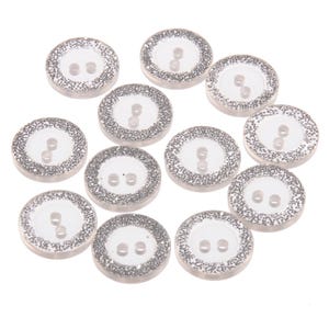 Peut inclure: Ensemble de boutons ronds transparents avec une bordure argentée scintillante. Chaque bouton comporte deux petits trous pour la couture. Les boutons sont disposés sur un fond blanc, pour les projets de couture et d'artisanat.