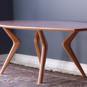 May include: A light wood coffee table with a long, oval-shaped top and four unique, angled legs. The table is sitting on a patterned rug.