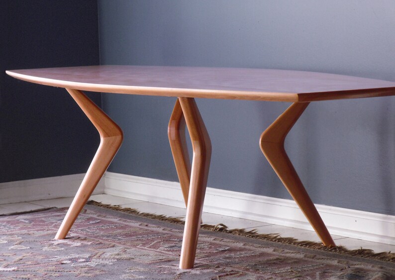 May include: A light wood coffee table with a long, oval-shaped top and four unique, angled legs. The table is sitting on a patterned rug.