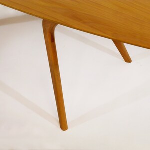 May include: Close-up of the legs of a wooden table. The table has a light brown finish and the legs are slender and tapered. The legs are angled slightly inward.