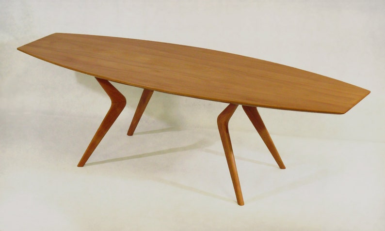 May include: A long, oval-shaped wooden coffee table with four unique, angled legs. The table is made of light brown wood and has a smooth, polished finish.