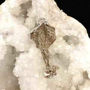 May include: A silver pendant necklace featuring a unique, textured design. The pendant has an organic shape with a detailed surface and a small flower at the bottom. The necklace chain is a delicate silver rope.