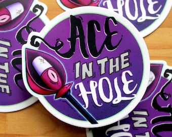 Puede incluir: Tres pegatinas redondas de color morado y verde con el texto "Ace in the Hole" en letras blancas y negras. Las pegatinas presentan una ilustración estilizada de una pica de carta de juego con un vendaje y una flor rosa.