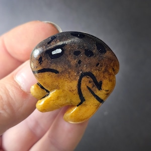 May include: A small, smooth, rounded stone painted to resemble a frog. The upper portion is a dark brown with black spots, while the lower section is a vibrant golden yellow. Black painted lines define the frog's eye, mouth, and folded leg structure.