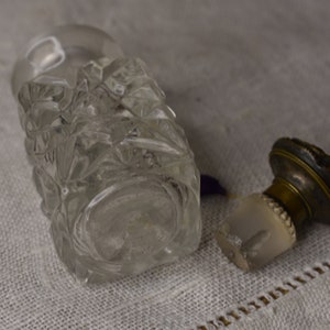 May include: A clear glass bottle with a faceted design and a brass stopper. The bottle is empty and has a vintage look.