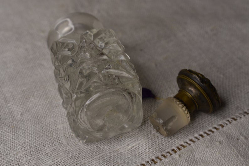 May include: A clear glass bottle with a faceted design and a brass stopper. The bottle is empty and has a vintage look.