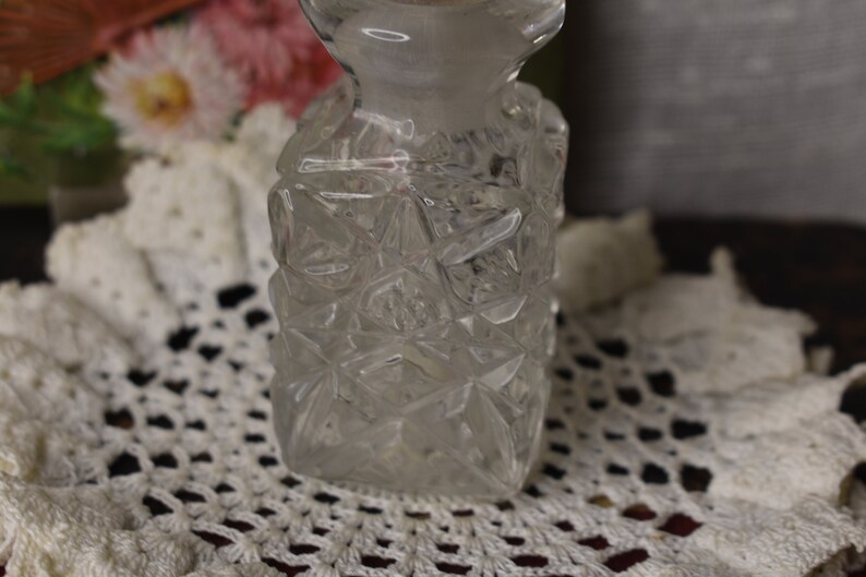 May include: A clear glass bottle with a diamond-shaped pattern. The bottle is sitting on a white crocheted doily.