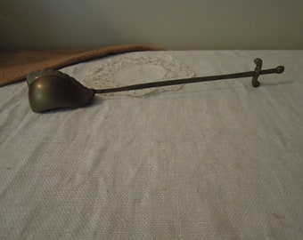 May include: A brass shoe horn with a sword-shaped handle. The shoe horn is curved and has a rounded tip. The handle is long and slender, with a cross-shaped guard and a pointed tip.