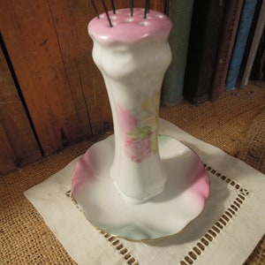 May include: A white porcelain needle holder with a pink and green floral design. The holder has a round base and a tall, slender neck with a pink top. The top has a series of holes for holding needles.