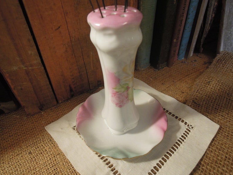 May include: A white porcelain needle holder with a pink and green floral design. The holder has a round base and a tall, slender neck with a pink top. The top has a series of holes for holding needles.