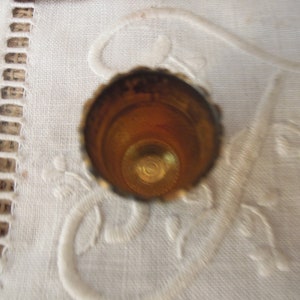 May include: A close-up shot of a vintage thimble. The thimble is made of gold-colored metal with a scalloped edge. The interior is a warm, golden hue. The thimble sits on a white, embroidered surface.