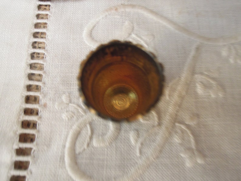 May include: A close-up shot of a vintage thimble. The thimble is made of gold-colored metal with a scalloped edge. The interior is a warm, golden hue. The thimble sits on a white, embroidered surface.
