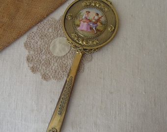 May include: A gold-toned hand mirror with a decorative handle and a round mirror face. The mirror face has a porcelain inset with a painted scene of two people in a garden.