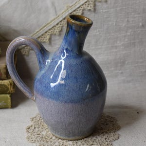 May include: A handmade ceramic pitcher with a blue and lavender glaze. The pitcher has a rounded body, a curved handle, and a spout with a brown rim. It sits on a small, beige doily, with a white ceramic rabbit and books in the background.