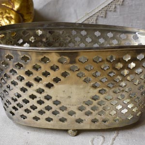 May include: An oval-shaped metal basket with a polished silver-tone finish. The sides feature a repeating lattice pattern of small, four-pointed cutouts. The basket is supported by small, rounded feet at the base, creating a raised structure.