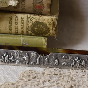 May include: An antique letter opener with a silver-toned handle featuring a detailed relief scene of people in a garden. The blade is a warm brown color. Books and lace are in the background.