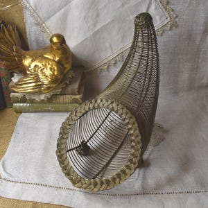 May include: A vintage-style, gold-toned metal cornucopia basket. The basket is shaped like a horn of plenty, with a woven metal design and a braided trim around the opening. A gold-colored bird statue is visible in the background.