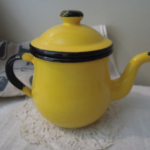 Puede incluir: Una tetera de esmalte amarillo con tapa y asa negras. La tetera tiene un cuerpo redondeado y un pico en el lateral.