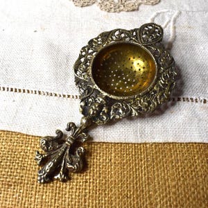 May include: A brass tea strainer featuring a circular bowl with small perforations, surrounded by an ornate, filigree-style border. The handle is shaped like a fleur-de-lis, and the metal surface displays a dark, aged patina with intricate relief details.