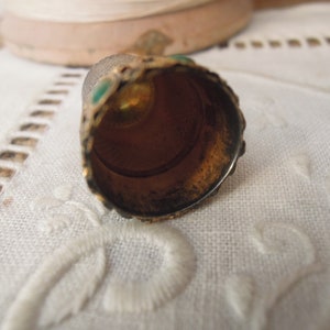 May include: Close-up of a vintage thimble with a gold-colored exterior and green accents. The thimble's interior is a warm, bronze color. The thimble is resting on a white, textured surface.