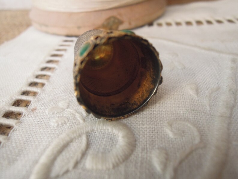 May include: Close-up of a vintage thimble with a gold-colored exterior and green accents. The thimble's interior is a warm, bronze color. The thimble is resting on a white, textured surface.