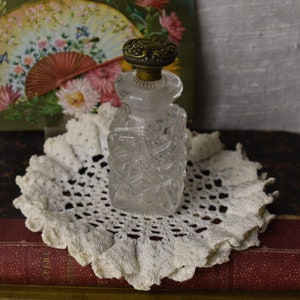 May include: A clear glass bottle with a textured surface and a gold metal top. The bottle is sitting on a white crocheted doily on top of a stack of old books.