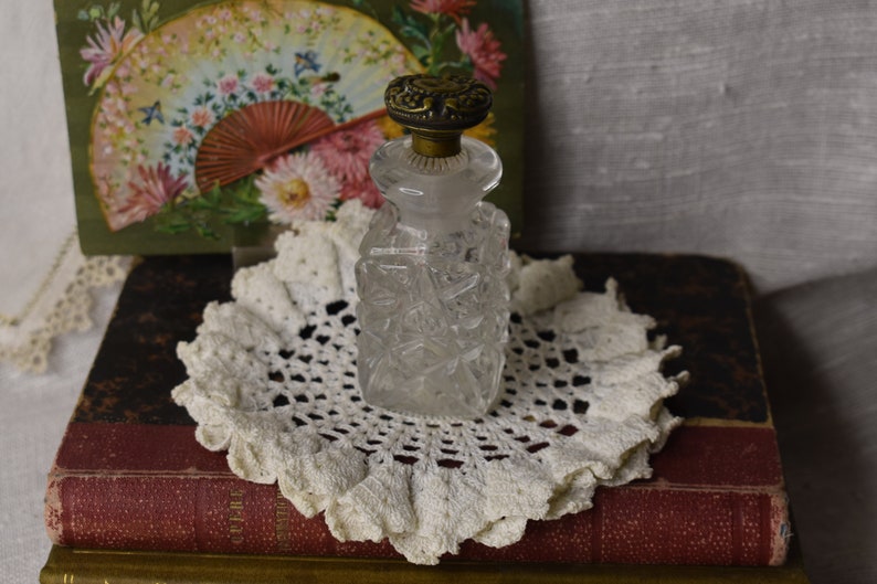 May include: A clear glass bottle with a textured surface and a gold metal top. The bottle is sitting on a white crocheted doily on top of a stack of old books.