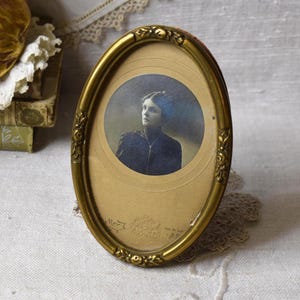 May include: An oval-shaped gold-toned metal picture frame featuring embossed floral details along the border. The frame contains a vintage sepia-toned portrait photograph of a person wearing a dark high-collared garment and a delicate necklace.