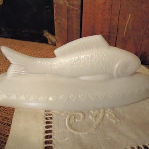 May include: A white milk glass butter dish lid shaped like a fish resting on an oval base. The fish features detailed scale textures and a dorsal fin, while the base is decorated with a repeating embossed heart pattern along the lower edge.