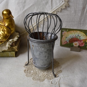 May include: A vintage metal container with a wire cage top. The container is gray and has three metal legs. A golden bird statue and a small floral print are in the background. The container sits on a lace doily.