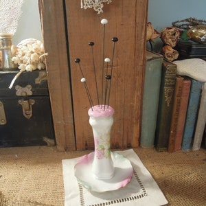 May include: A white porcelain pincushion with a pink floral design and black and white pins. The pincushion is on a white saucer with a pink and green rim.