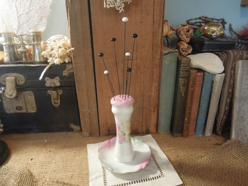 May include: A white porcelain pincushion with a pink floral design and black and white pins. The pincushion is on a white saucer with a pink and green rim.