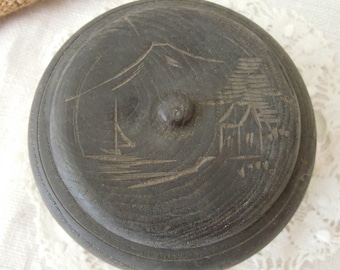 Op de afbeelding: Een donkere houten doos met een gegraveerd ontwerp van een zeilboot en een huis op een heuvel. De doos heeft een knop aan de bovenkant.