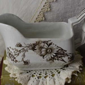 Könnte beinhalten: Weiße Keramik-Soßenkännchen mit braunem Blumendesign. Die Kanne hat einen Griff und steht auf einer Spitzenunterlage.