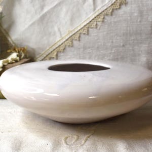 May include: A low, oval-shaped, off-white ceramic vase with a dark interior. The vase has a smooth, glossy finish and a wide, circular opening. The background includes a small potted plant and vintage books.
