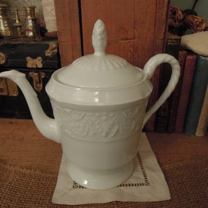 May include: A white porcelain teapot with a lid, spout, and handle. The teapot has a decorative embossed floral pattern around the middle and sits on a small white square doily. The teapot is a classic design.