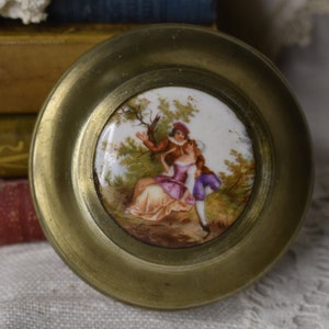 May include: A brass trinket box with a round, porcelain lid featuring a painted scene of a couple in a garden setting. The couple are dressed in 18th-century clothing and the woman is wearing a purple dress.
