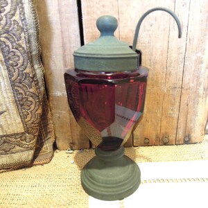 May include: A vintage ruby red glass jar with a dark green lid and base. The jar has a unique shape with a curved handle attached to the lid. The jar is likely used for storage or display.