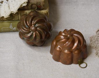 Puede incluir: Dos moldes antiguos de cobre con diseño floral. Los moldes tienen un patrón en espiral y un pequeño anillo de latón para colgar. Se colocan sobre una superficie de lino, con libros antiguos y encaje en el fondo.