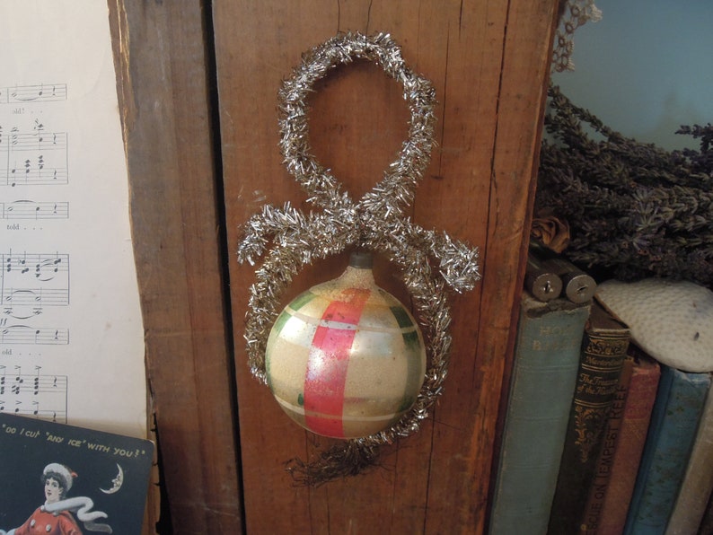 May include: A vintage glass Christmas ornament with red, green, and white stripes. The ornament is decorated with silver tinsel.