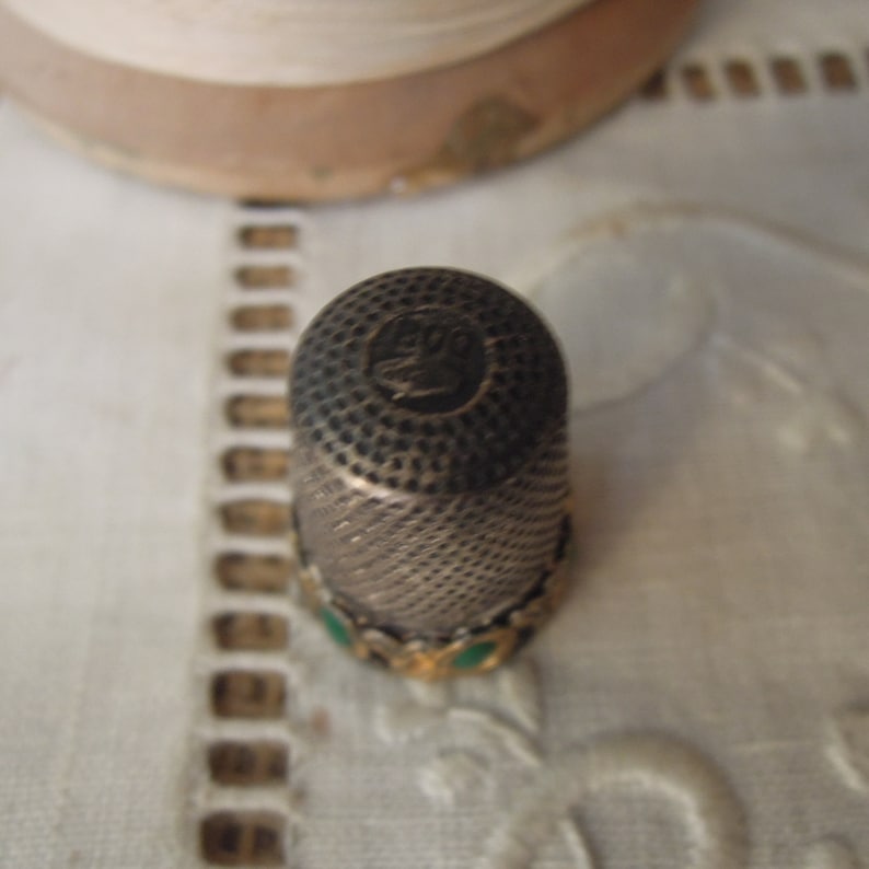 May include: A vintage silver thimble with a textured surface and a decorative band with green accents. The thimble has a dark top with an embossed design. A sewing tool for crafting and needlework.