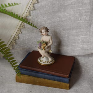 May include: A porcelain figurine of a cherub holding a bunch of grapes, atop a stack of antique books. The cherub is nude with detailed facial features and is seated on a decorative base with gold trim. The books are brown, blue, and gold.