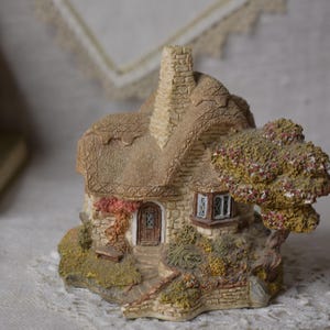 May include: A miniature cottage figurine with a thatched roof, stone walls, and a small tree. The cottage has a door, windows, and a stone chimney. The figurine is made of a light brown and gray material.